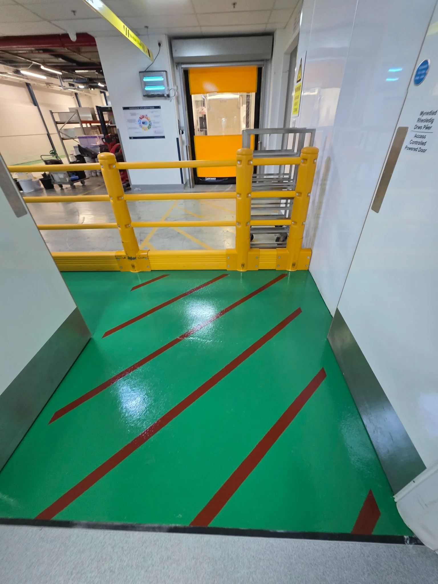 Red hatched safety exclusion markings on a green industrial floor beside protective barriers